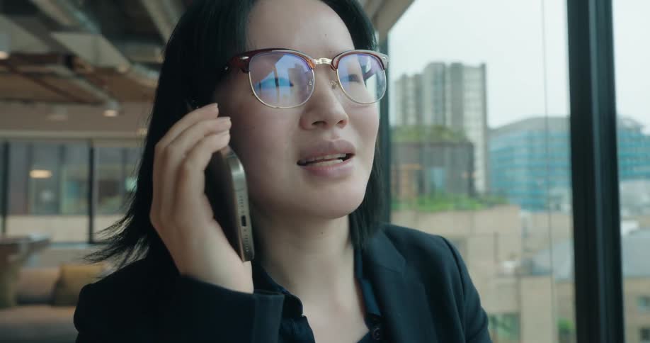 Looking forward an asian woman in a dark business suit and glasses is focused while speaking on a cell phone inside a modern office with a city skyline visible through the window