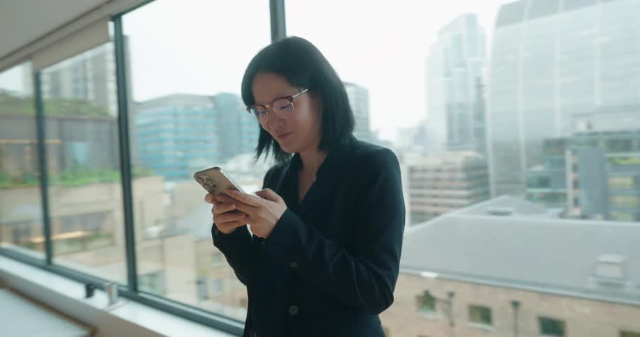 A woman standing next to a window looks down at her smart phone