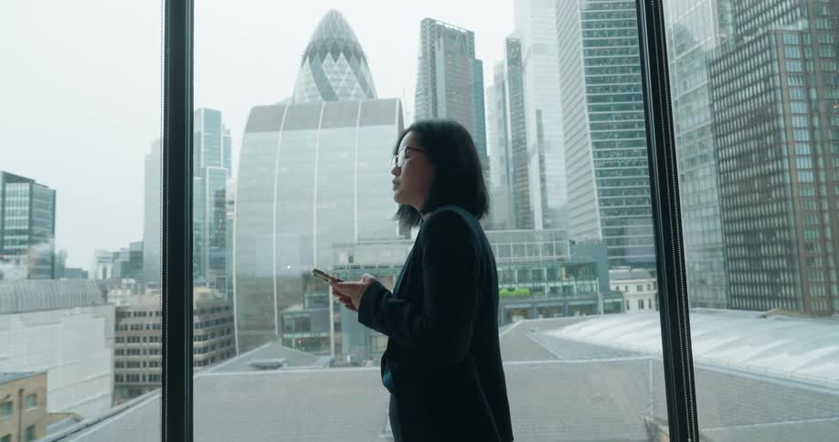 A woman stands by a window checking her phone in an office