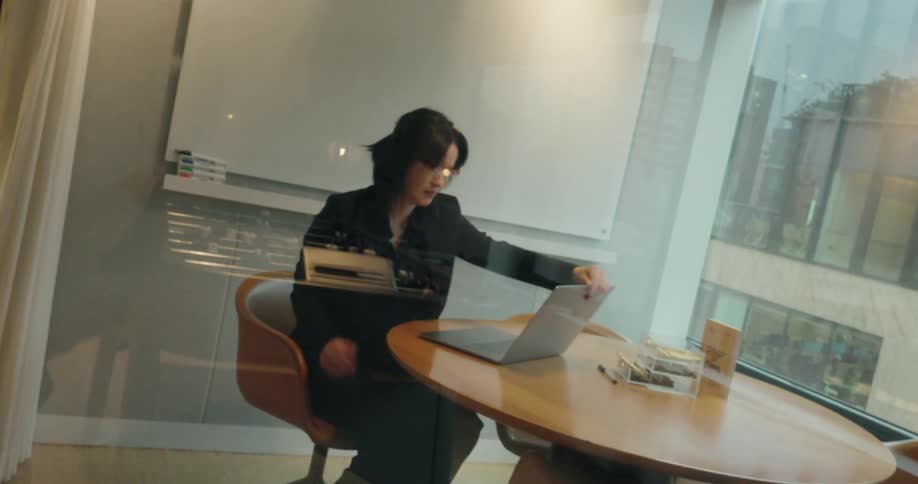 A woman in an office walks to a room that has a table and begins to type on a laptop