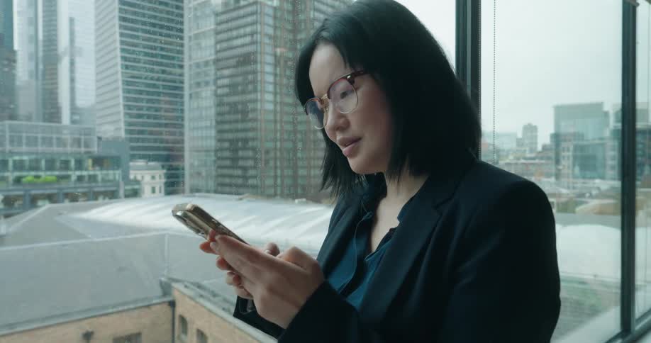 The video shows a woman dressed in professional attire a dark blazer standing indoors next to a large window overlooking a city skyline of tall buildings she is holding a goldcolored smartphone and initially appears focused on texting or browsing