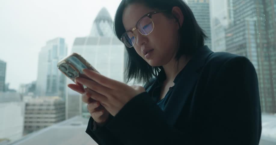 An asian woman wearing glasses stands beside a window operating her smartphone with the city skyline visible through the glass