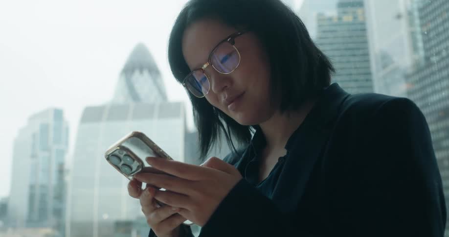 Young asian woman with glasses wearing black formal clothing texting on a smartphone next to a window in a metropolitan area outdoors during day time