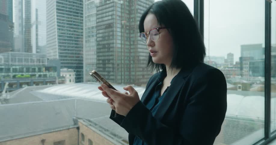 A professionallooking woman in a black suit and glasses is looking out a large window at a city viewshe is holding and looking at a phone then puts it down and smiles slightly as she looks toward the skyscrapers outside