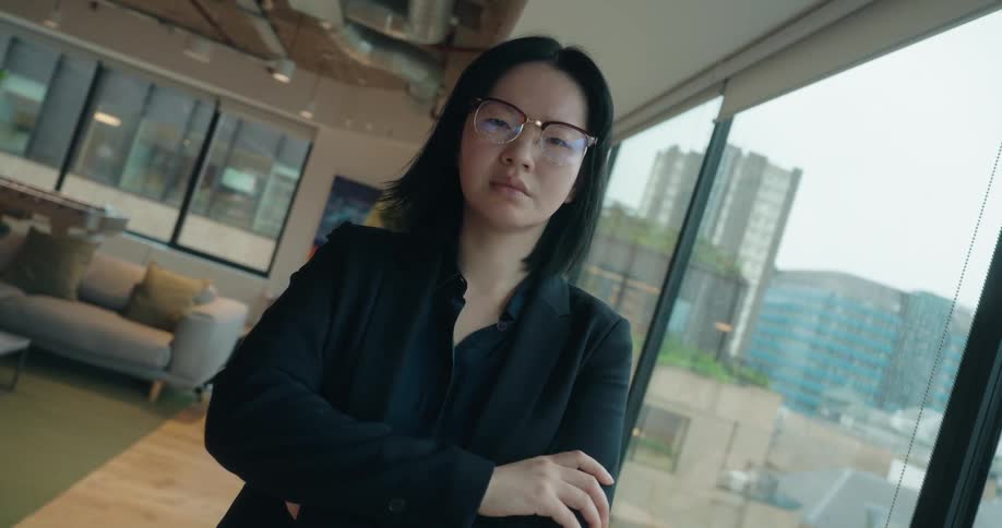 A young asian woman wearing a black blazer suit and glasses stands inside a highrise building beside the window