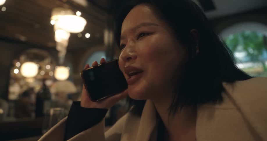 A closeup shot focuses on a woman as she holds a mobile phone to her ear and talks seated indoors in a lowlit elegant restaurant with large hanging light fixtures in the background