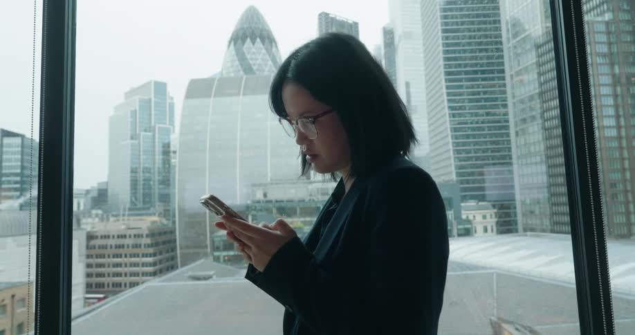 An asian woman wearing glasses and a suit stands inside a highrise building beside a window typing on her smartphone with the city skyline visible through the glass
