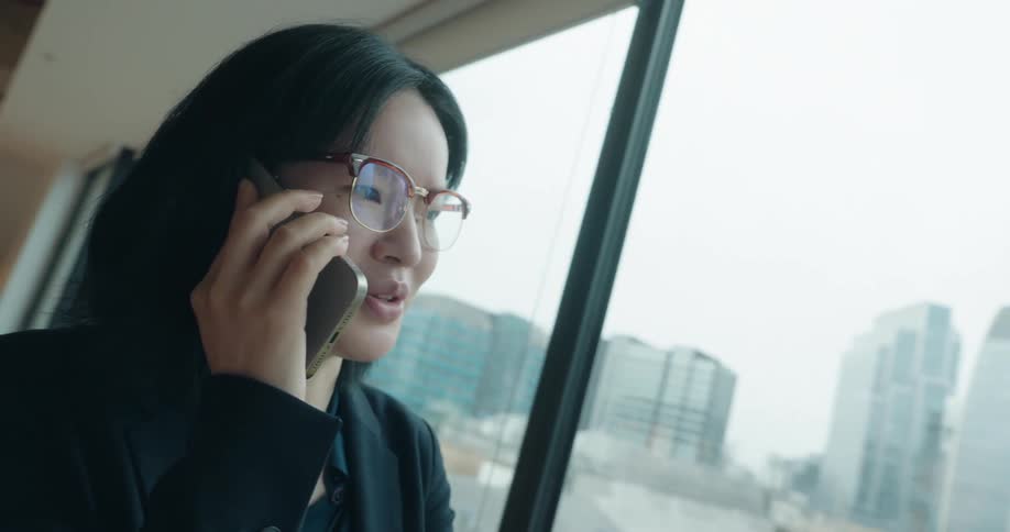 The video features a closeup of a person with short dark hair and eyeglasses dressed in a black blazer speaking on a cell phone they are standing indoors near a large window with a backdrop of city buildings visible outsidethe person appears enga