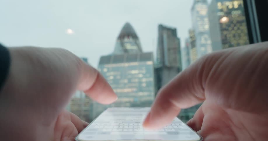A person is texting on their phone as they face the window looking out into the city