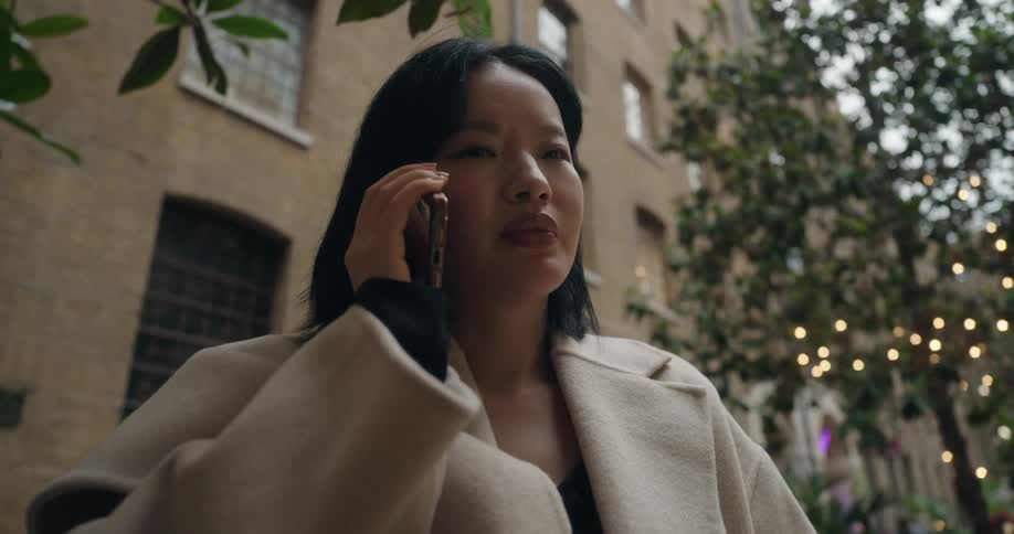 A young woman with dark hair and a beige coat talks on a mobile phone on an urban street lined with brick buildings trees and string lights