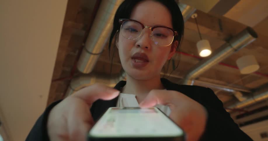 This is a lowangle closeup shot of a young woman wearing glasses and business attire intensely focused on typing on a smartphone she holds directly toward the camerathe background shows exposed ductwork and modern pendant lighting suggesting she i