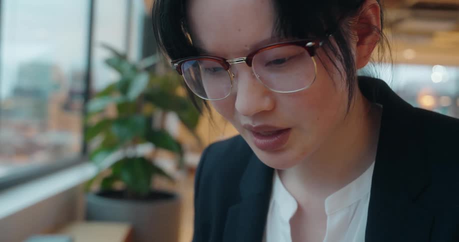 A young businesswoman in a black blazer and glasses works on a laptop by a large window overlooking a city skyline