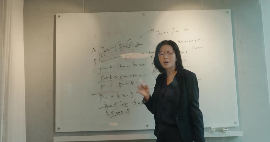 A woman in a black blazer and glasses stands in front of a glass whiteboard covered in handwritten notes and diagrams actively gesturing and speaking as if explaining a complex topic in a business presentation