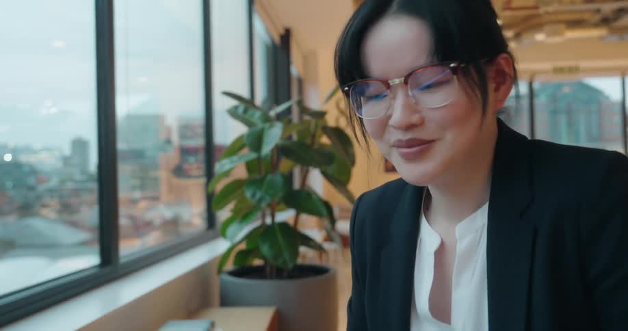 A young woman in professional attire and glasses sits near a large window overlooking a city and focuses intently on a laptop resting on her lap then smiles slightly while working in a bright modern office lounge area