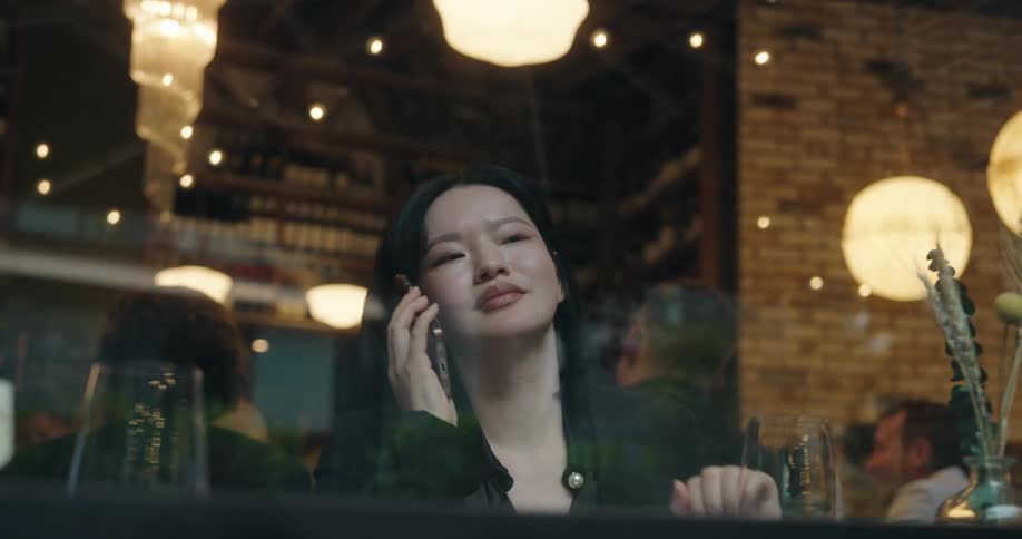 Woman talks on phone in cozy restaurant seen through window with reflections and bottles in the background