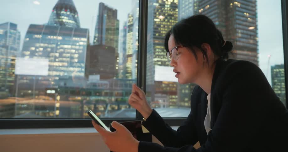 A woman wearing a blazer and glasses looks at her smartphone by a large window with a dramatic view of a city skyline lit up at night
