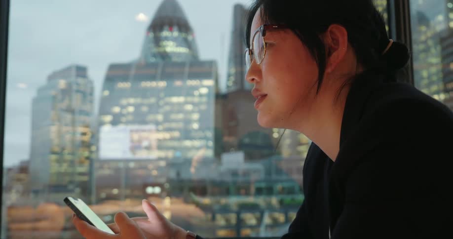 The video shows a closeup of a person wearing glasses and a dark blazer looking intently down at a smartphone they are holding in their hands they have their hair pulled back and are wearing a wristwatch
