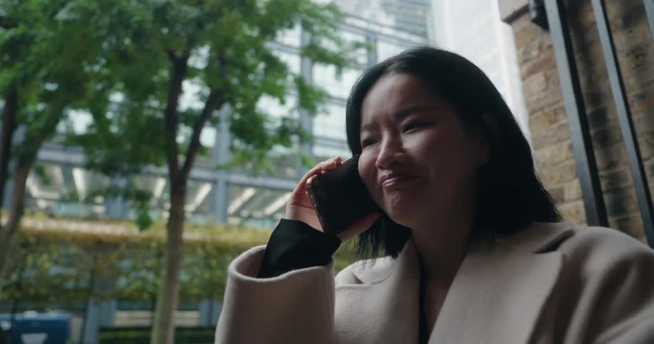 A woman with dark hair and wearing a lightcolored coat is standing outdoors in a modern urban environment engaged in a phone conversation she smiles as she speaks with a glass skyscraper and trees visible in the background
