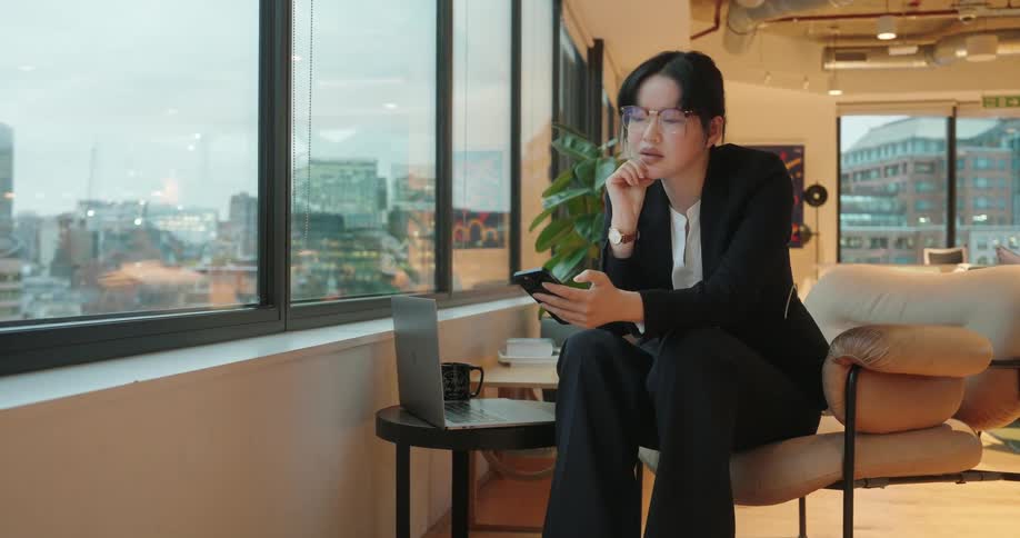 Person gazes at city through window in modern office holding phone beside table with laptop