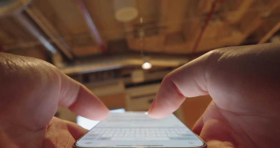 A persons hands holding a smartphone and rapidly using both index fingers to type text onto the bright screen in an interior space with exposed wooden beams and warm overhead lighting