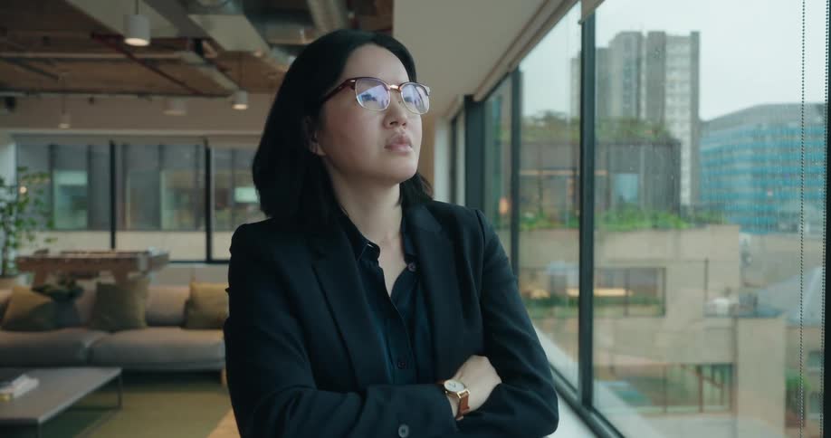 A professional woman in a suit stands by a large office window looking out at the city skyline she pauses to put on her glasses folding her arms and taking on a focused thoughtful expression as the camera zooms in