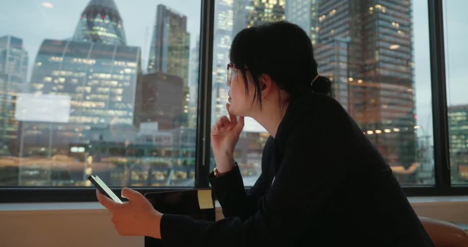A lady seated indoor near a glass window typing on a smartphone at sunrise