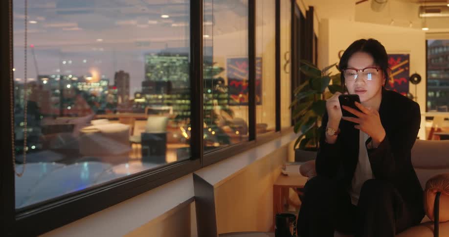 Young asian woman wearing glases and formal clothing sitting and texting on a smartphone next to a window showing a metropolitan city outdoors during dusk or dawn