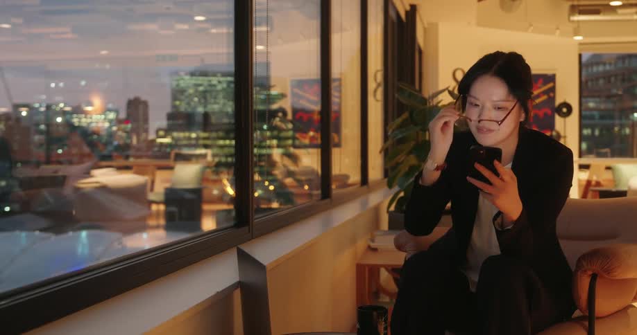 Woman seated in a nightlit office looks at her phone near the window