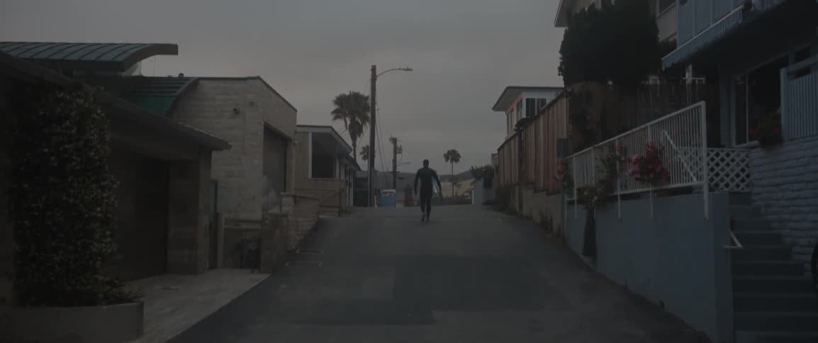 Man holding surfboard walking down a street at dusk