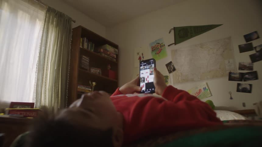 Young male person lying down in bed holding a smartphone on his hands and lifting it up and down over his chest indoors during day time