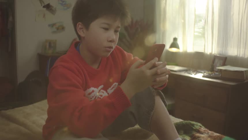 A young boy in a red sweater and khaki shorts sits on a bed happily using a red smartphone in a bright sunlit room with trophies and drawings