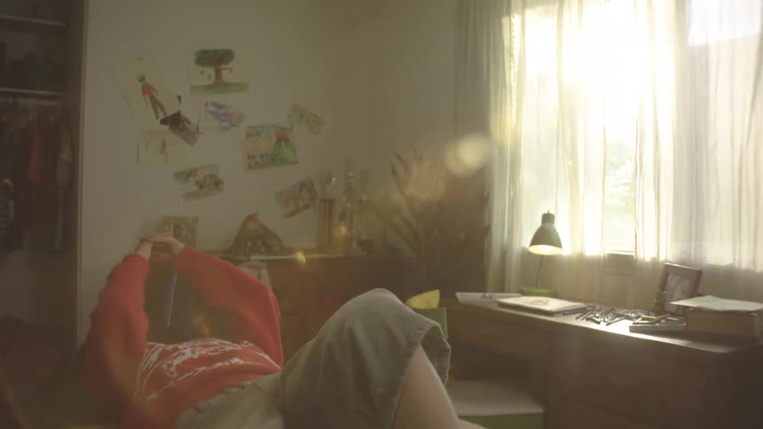 A low angle shot of a teenage boy lying in bed with with a phone in his hands in a bright sunlit room decorated with childhood drawings and a desk
