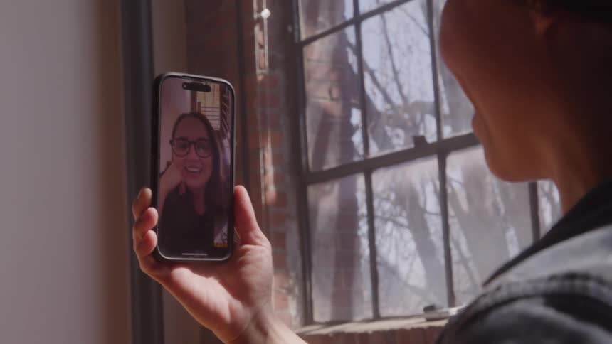 A person holding a smartphone during a video call where the screen displays a smiling woman wearing glasses and the person holding the phone raises their hand in a brief waving gesture