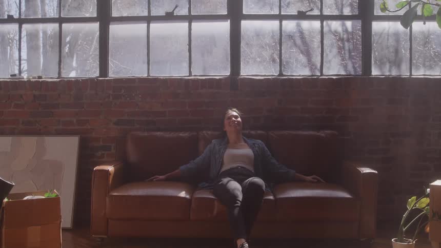The video shows a woman walking towards a brown leather sofa in a sunlit room with large industrialstyle windows and a brick wall she then excitedly jumps or collapses onto the sofa settling back and appearing relaxed