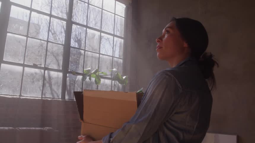 Woman smiles while holding a cardboard box filled with items near a bright window