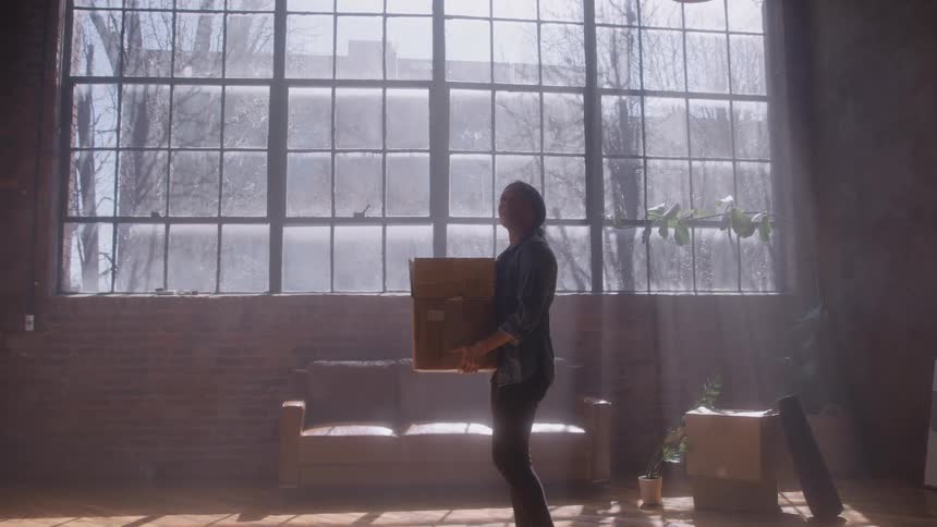 A woman wearing dark casual clothes walks into a large sunlit loft space carrying a cardboard box before she stops to look around the room with exposed brick walls and a massive grid window dominating the background