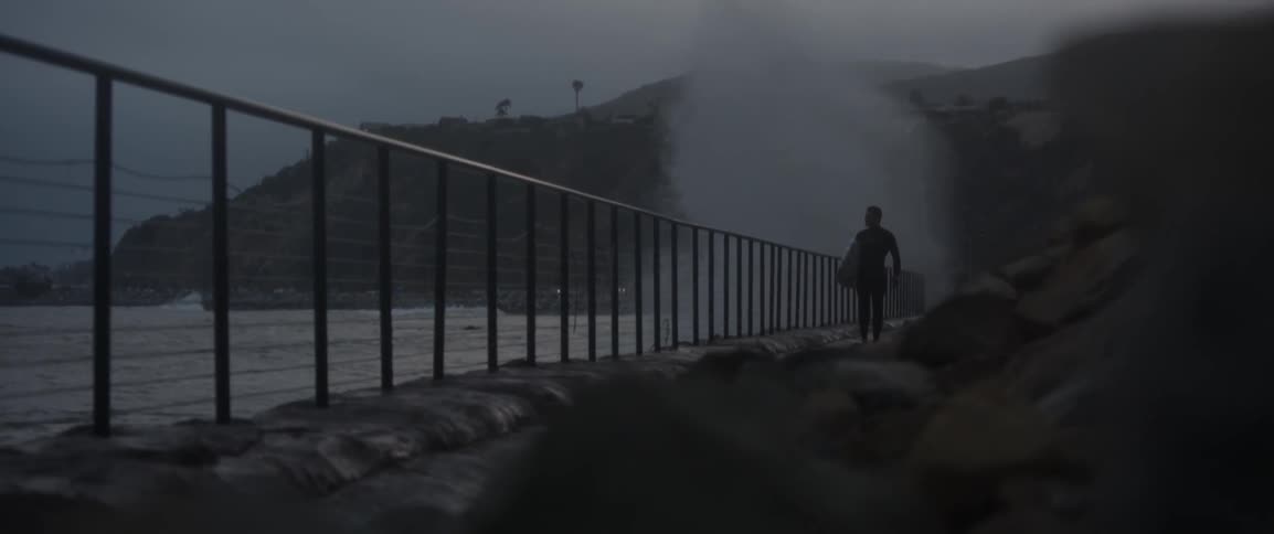 A man carries a surfboard while walking along a fence near the ocean at night