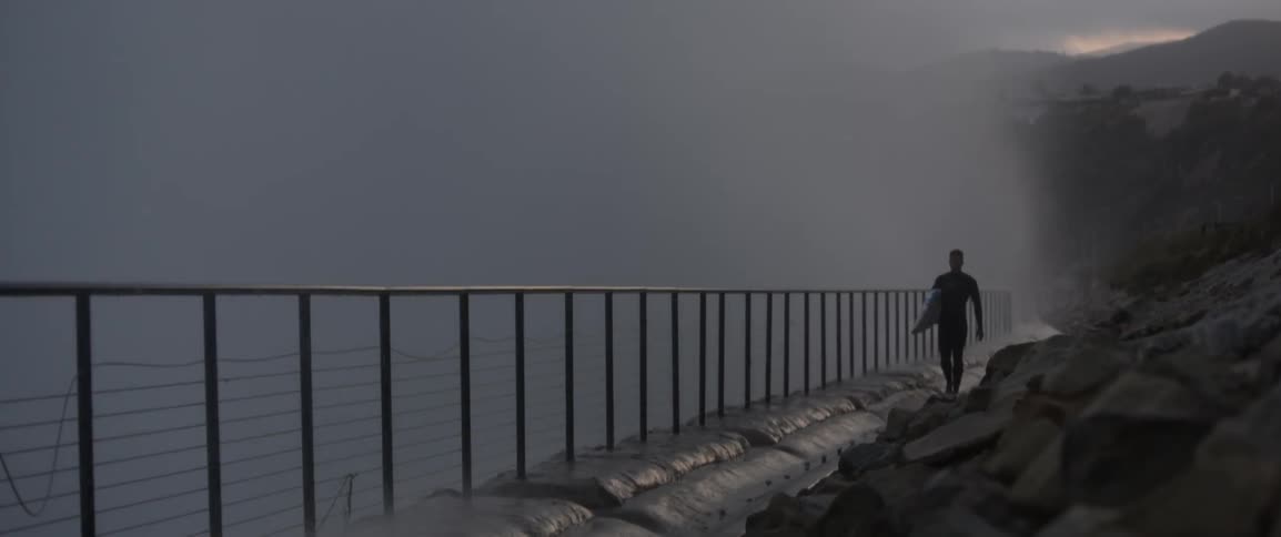 Waves splashing high against a retaining wall as a surfer walks by carrying his board