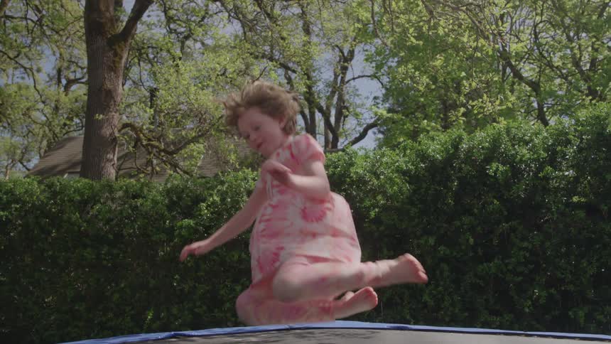 A barefoot white girl in a pink floral outfit jumps high on a trampoline with an energetic emotion in a sunny backyard setting