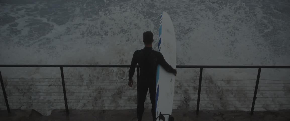 Man holding surfboard standing on walkway watching crashing waves