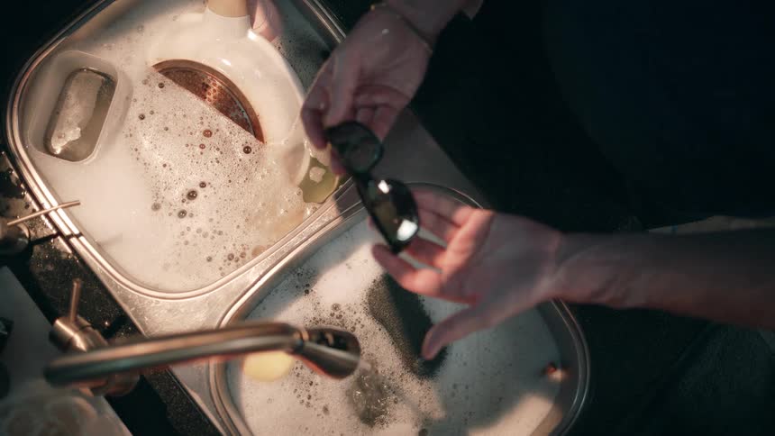A couple is in the kitchen washing dishes at the sink when the man picks up a pair of sunglasses from the sink