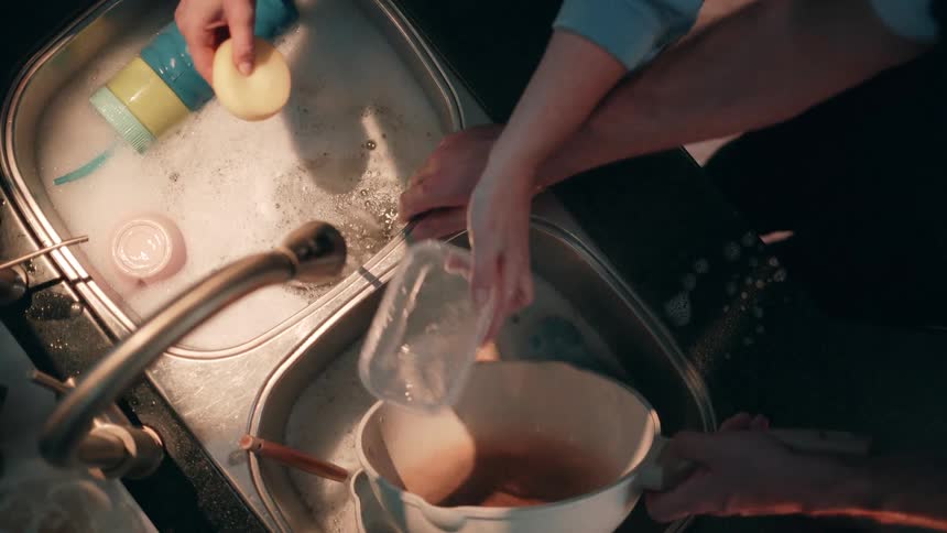 The video is a topdown view of two people washing dishes in a doublebasin kitchen sinkone person is washing items with a sponge in the bubbly basin while the other holds a saucepan full of liquid over the second basin and uses a small item to scrap