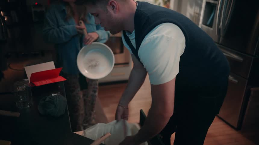 A man in a black vest and white shirt takes out the trash while a woman in a denim jacket and yellow headphones cleans up dishes in a modern kitchen