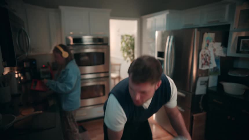 A man cleans up in a modern white kitchen while a woman in the background wears headphones and prepares coffee on a dark morning