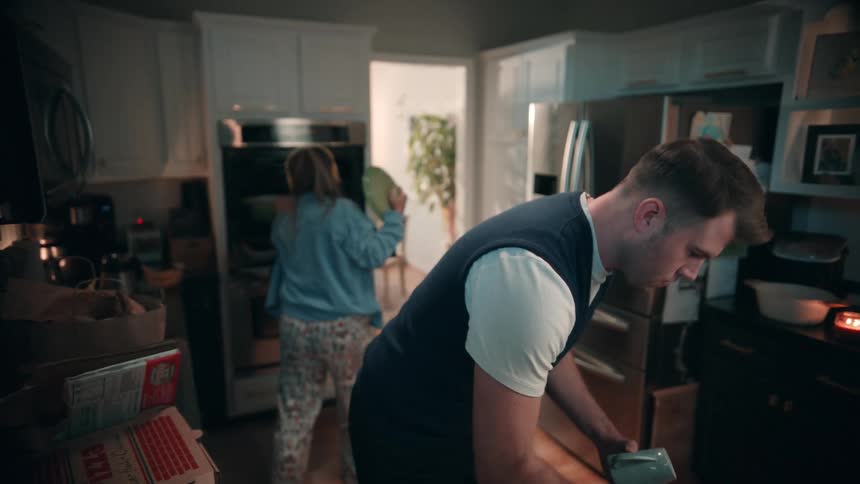 A man in a white tshirt and dark vest is shown in the foreground bending down to tidy up some items on a lower cabinet in a kitchenin the background a woman wearing headphones and pajamas is standing by an oven or microwave seemingly busy with a ta