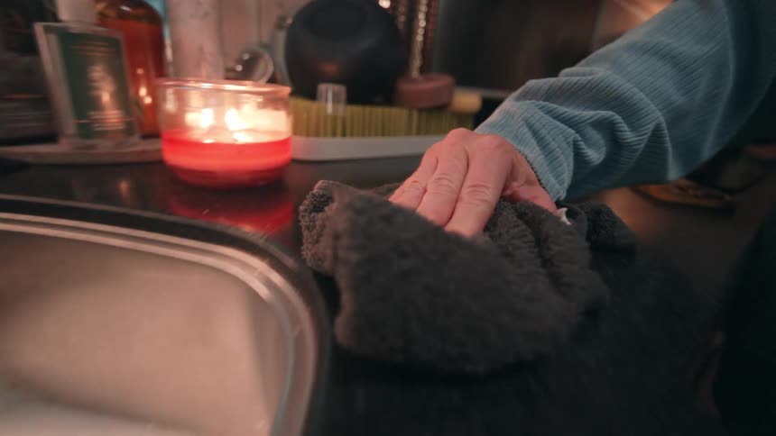 A womans hand wearing a blue sleeve actively wipes a dark reflective kitchen countertop using a dark grey terry cloth near a stainless steel sink with a glowing red candle visible in the background