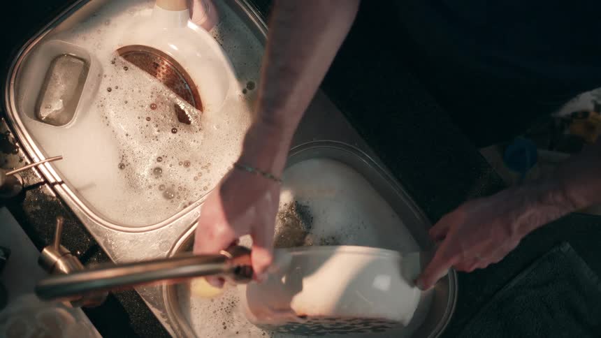 An overhead shot shows a persons hands actively submerged in a doublebasin kitchen sink filled with soapy water scrubbing a large white cooking pan with a handle and rinsing a green patterned dish