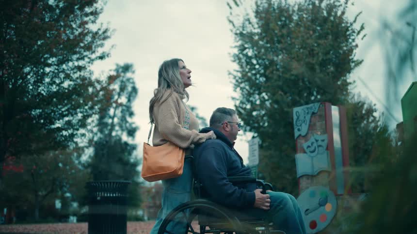 A caring woman in a beige sweater and jeans stands next to a man in a wheelchair outdoors in a park surrounded by trees