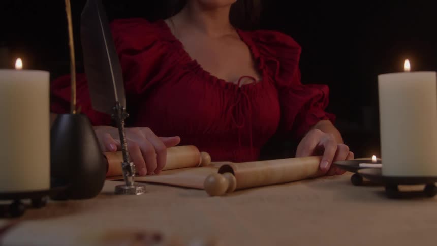 A woman in a historical red dress is intently unrolling a scroll by candlelight suggesting a scholarly task