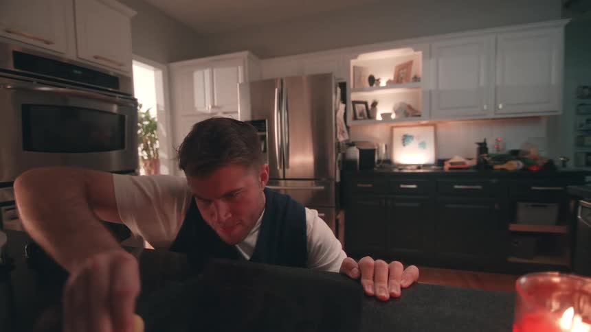 A man wearing a white tshirt and dark vest leans over a black countertop in a contemporary kitchen actively wiping the surface with a small cloth or sponge before standing upright and turning away from the counter